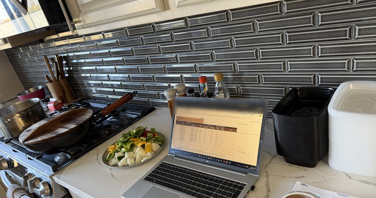 A Settel cook day: the batch log open on a laptop between the stove, prepped vegetables, containers and labels